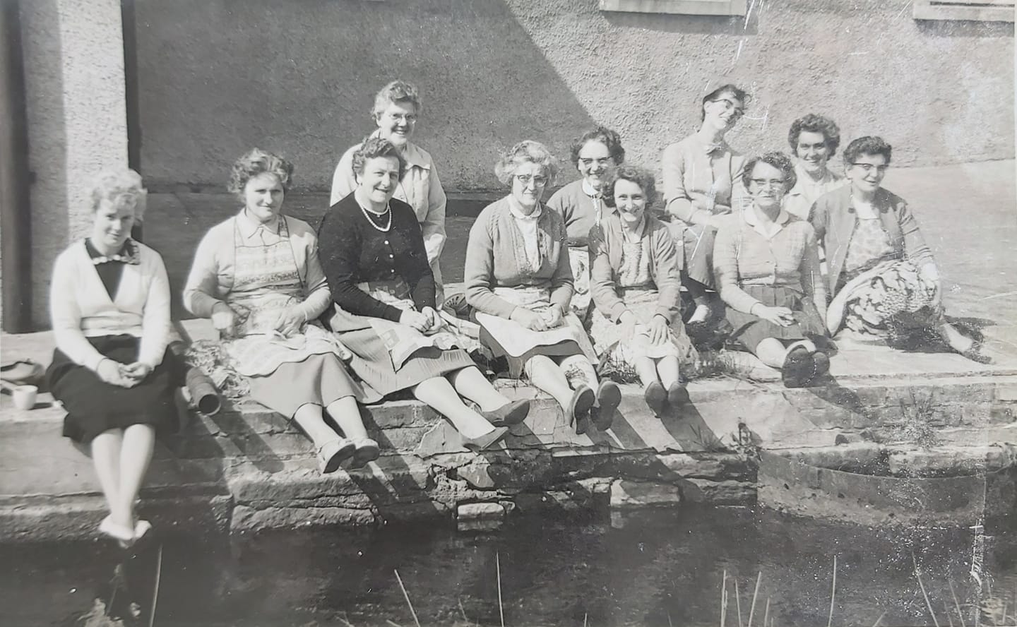 A black and white photo from the 1960s of a group of women workers at the Millers Factory in Cockermouth sitting on the side of the mill race in the sunshine