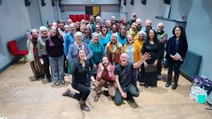 The community choir in the auditorium of the Kirkgate Centre. Jessie McMeekin, Jordan Worsfold and Dave Camlinn are kneeling down at the front, singing and smiling.