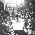 VE Day party for evacuees in Cocktons Yard, Cockermouth, 1945 A black and white photo from the 1940s with a large group of women holding babies and older children sitting round a table outdoors in an enclosed yard. There are flowers on the table and bunting hanging from the buildings.